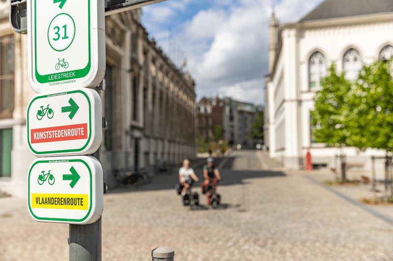 Wegbeschreibung für eine Fahrradroute in Brügge auf einer Stange mit Radfahrern im Hintergrund